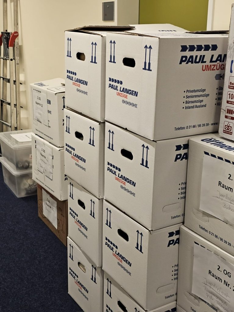 Gestapelte weiße Umzugskartons der Firma Paul Langen mit Tragegriffen und Aufrichtungspfeilen in einem Büroraum