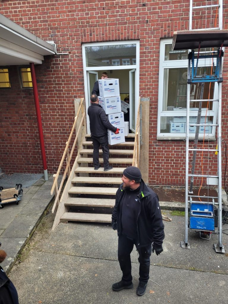 Umzugsteam trägt Kartons über Holztreppe in Backsteingebäude, Mitarbeiter koordiniert Transport vom Boden aus