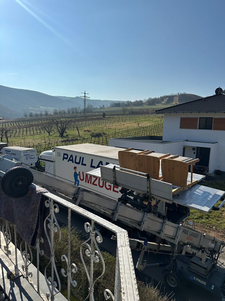 Umzugswagen von Paul Langen transportiert Kartons vor Wohnhaus in ländlicher Weinbauregion mit Hügeln im Hintergrund