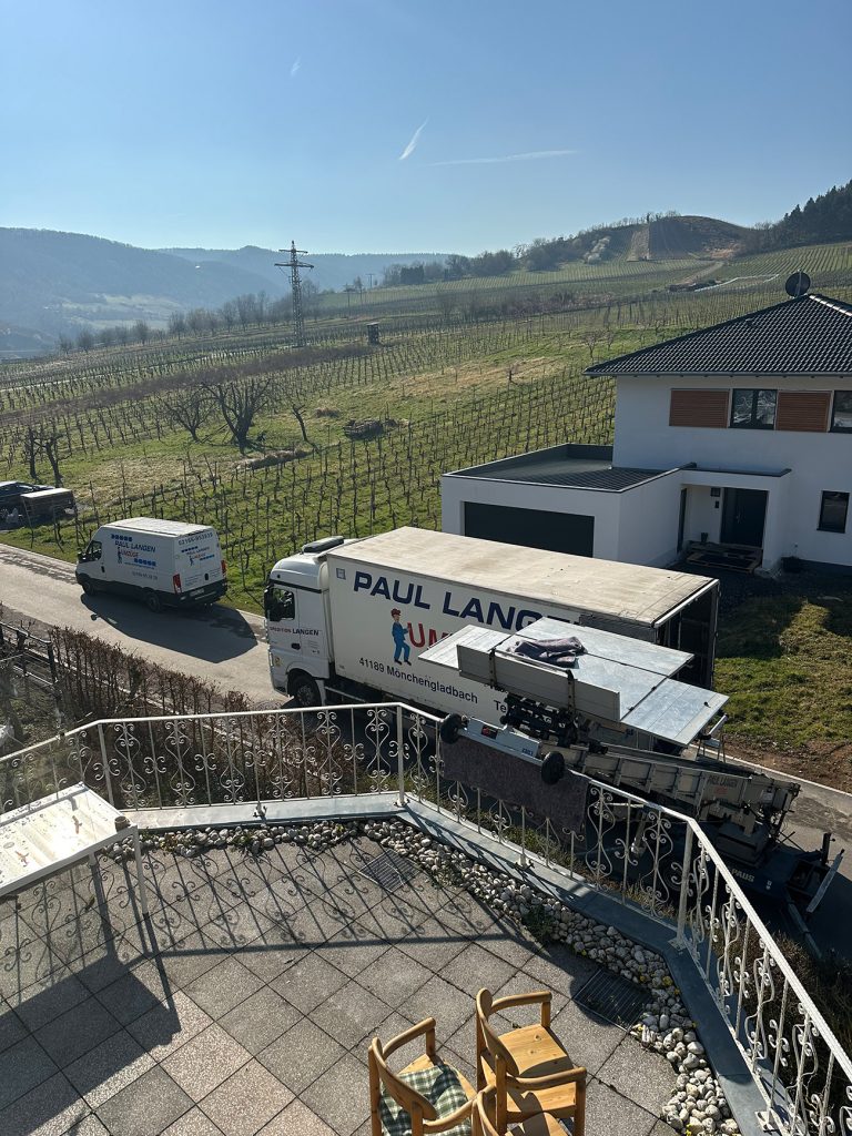 Umzugswagen von Paul Langen vor modernem Haus in Weinberglandschaft mit Terrasse und Balkongeländer